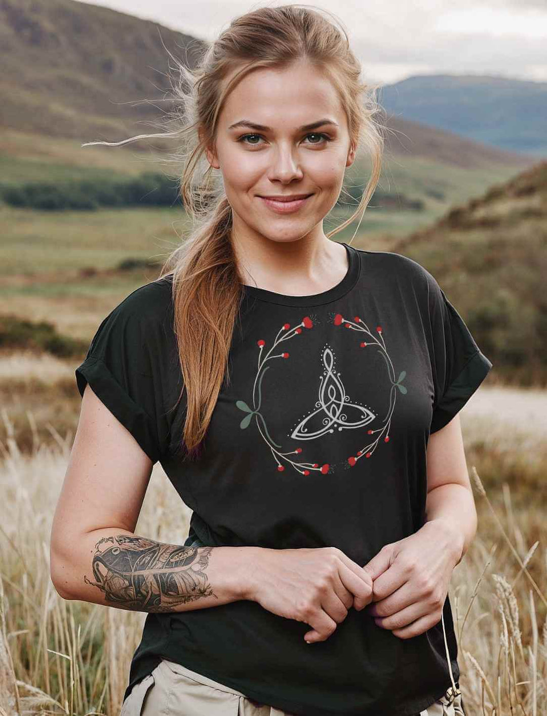 Eine junge Frau mit blondem Zopf steht vor einer mystischen Landschaft und trägt das Celtic Mother Extended Shoulder Tee in Schwarz. Das keltische Muttersymbol leuchtet auf dem Stoff, eingerahmt von filigranen Naturverzierungen – ein Zeichen der ewigen Verbundenheit.