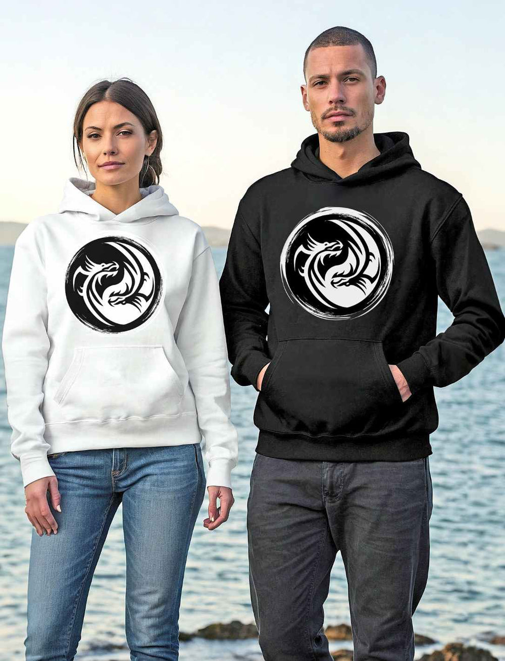Ein Paar trägt 'Drachensiegel des Gleichgewichts' Unisex Hoodies in Weiß und Schwarz am Ufer, das Design zeigt ein eindrucksvolles Yin-Yang-Drachenmotiv.