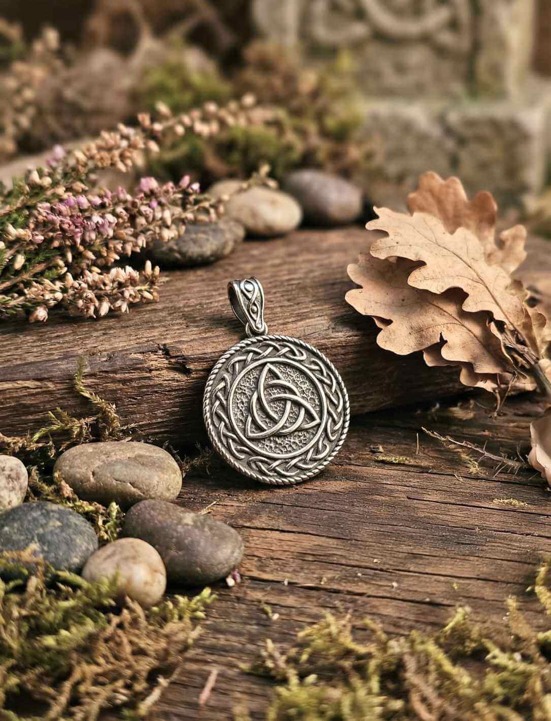 Keltischer Triquetra Anhänger mit Flechtrahmen, liegend auf Holz mit Steinen, Moos und Herbstblättern