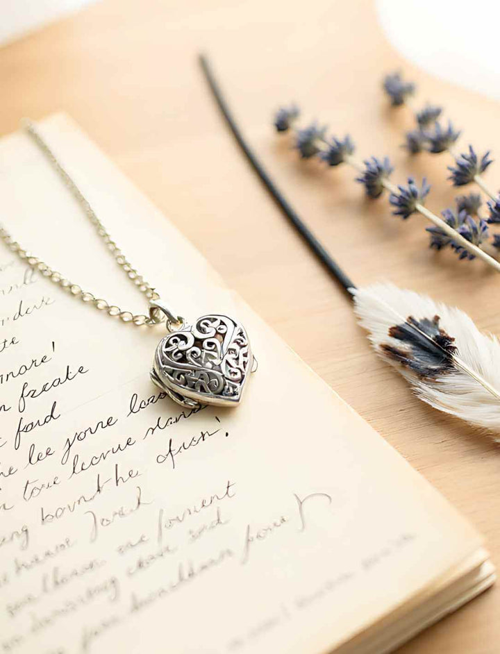 Silberanhänger in Herzform mit Ornamenten und kleinem Geheimfach, eingefasst an einer schwarzen Lederkette. Im Hintergrund alte Blätter und Sonnenlicht, eine Hand greift nach dem Schmuckstück – mystisch und authentisch in Szene gesetzt.
