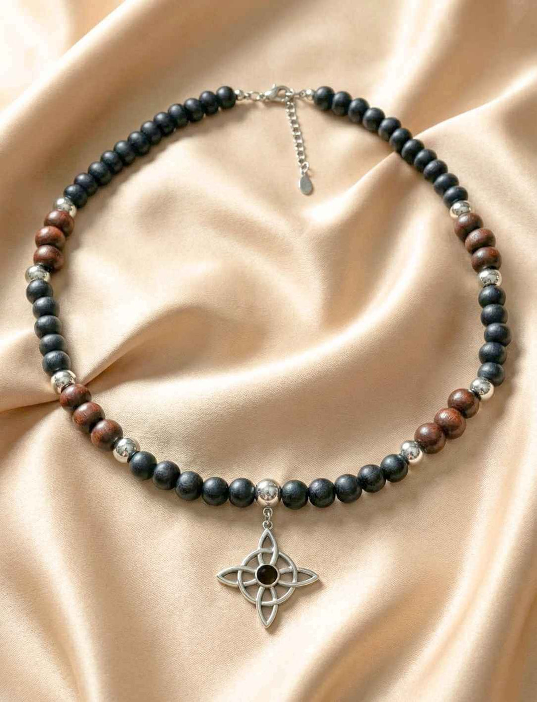 Holzperlenkette mit silberfarbenem vierarmigem Knotenanhänger und Onyxperlen auf Satin Hintergrund.