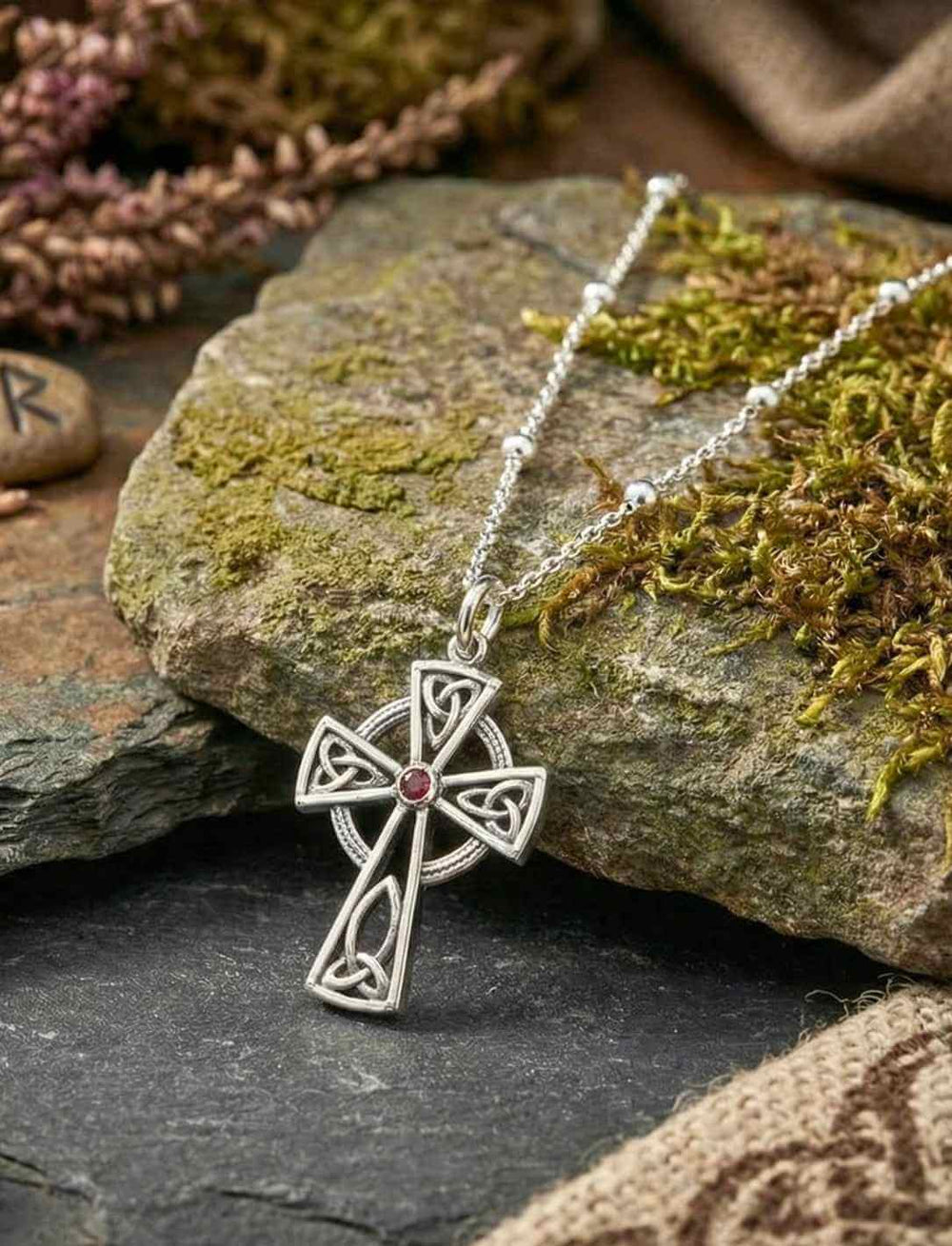 Keltisches Kreuz aus Silber mit rotem Stein auf Holz und Moos an Silberkette.