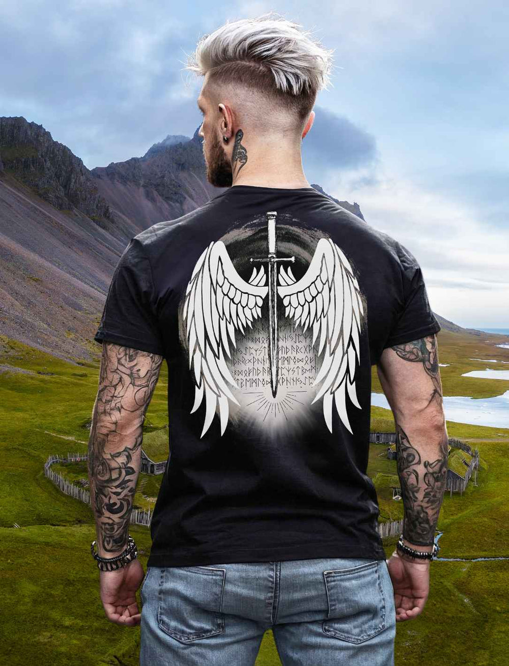 Junger Mann im Viking-Stil trägt ein schwarzes T-Shirt und blickt von einem Hügel auf die atemberaubende nordische Landschaft hinunter.