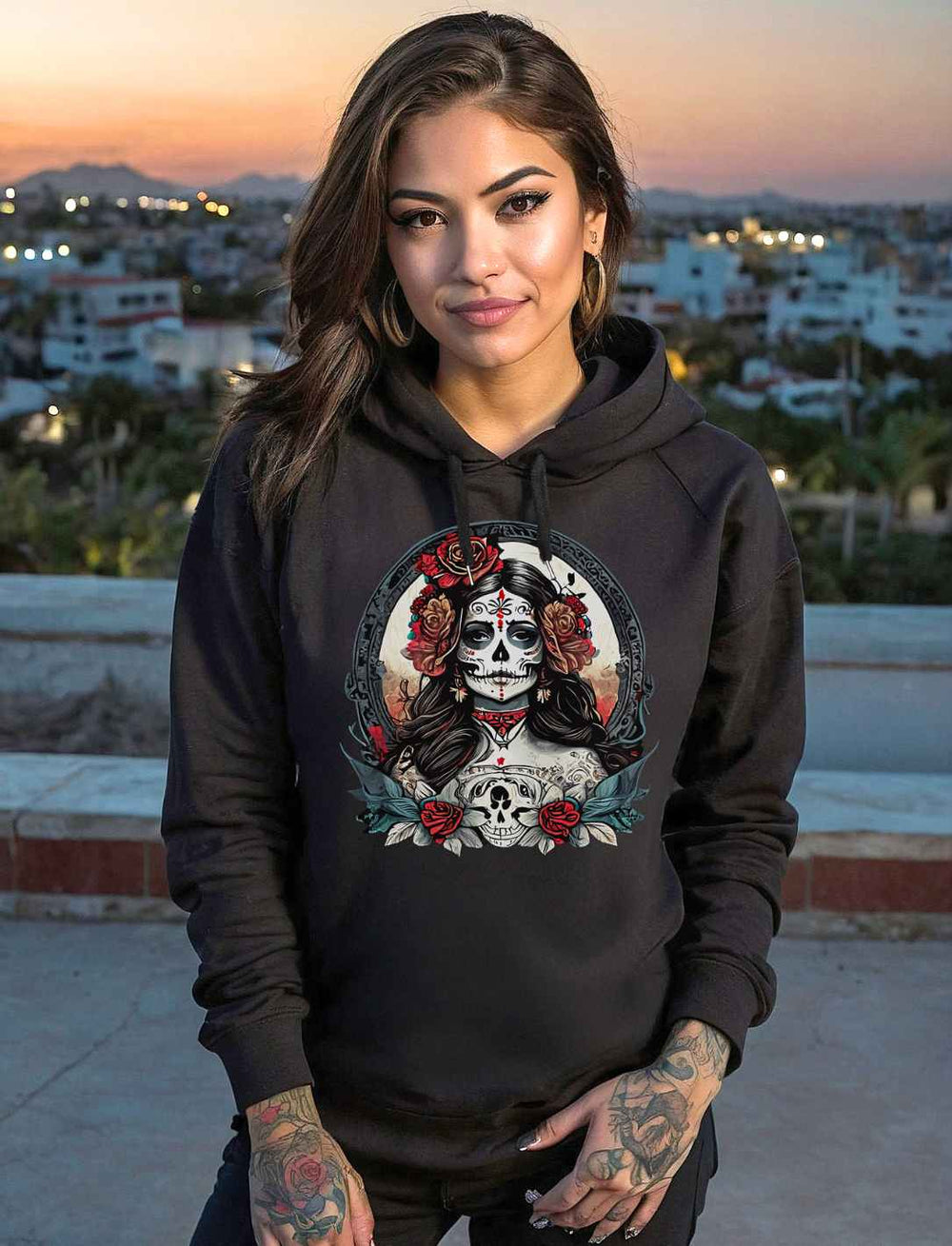 Frau trägt einen schwarzen La Catrina Damen Organic Hoodie mit farbenfrohem Día de los Muertos-Design, fotografiert bei Sonnenuntergang auf einer Terrasse mit Stadtblick im Hintergrund.