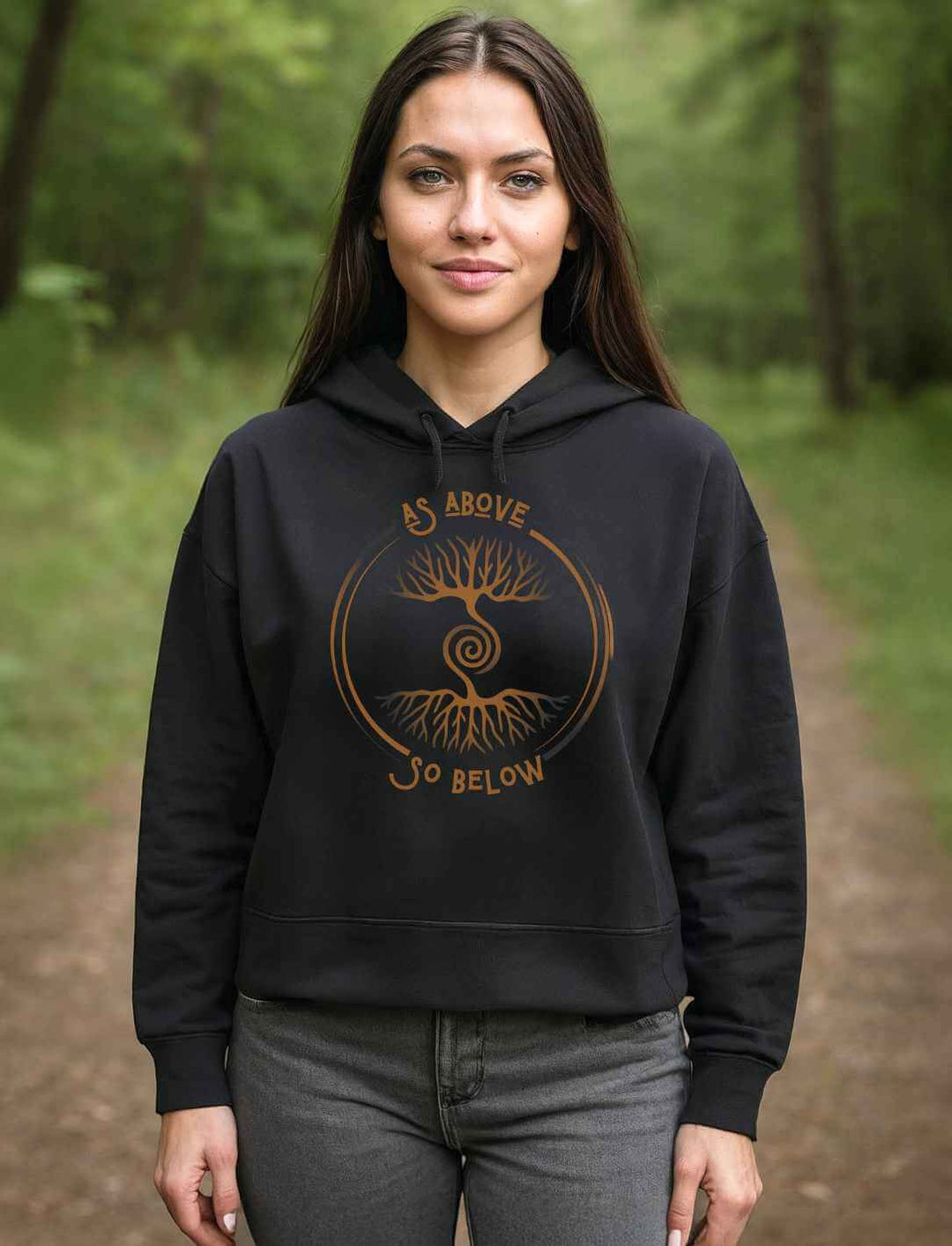 Frau trägt schwarzen Damen Organic Boxcut Hoodie „Lebenswurzel der Verbundenheit“ mit kupferfarbenem Weltenbaum-Motiv im Wald