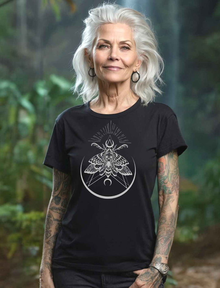 Ältere Frau mit grauem Haar trägt das “Lichtbringer der Verwandlung” Damen T-Shirt in Schwarz – Mystisches Design mit Nachtfalter-Symbolik, fotografiert in einem natürlichen, grünen Außenbereich.