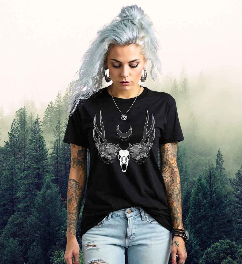 Ein weibliches Model mit eisblauem Haar trägt ein schwarzes "Lunas Mondgeweih" T-Shirt vor einem mystischen Waldhintergrund, die Ästhetik ergänzt durch ihre sichtbaren Arm-Tattoos.