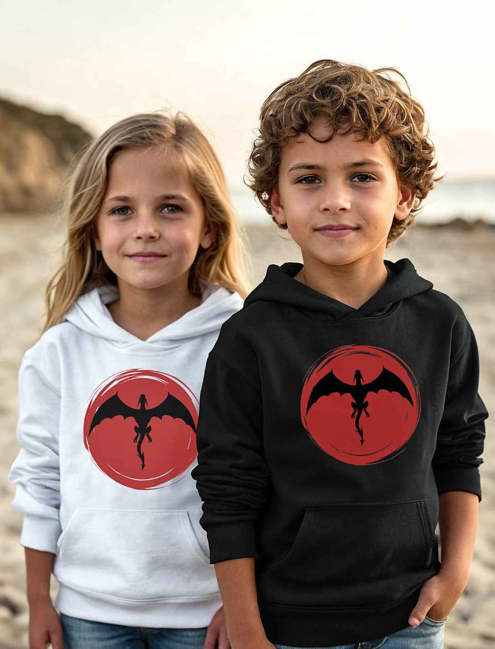Zwei Kinder am Strand, das Mädchen in einem weißen und der Junge in einem schwarzen 'Saga of the Dragon' Bio-Hoodie, lächeln in die Kamera und präsentieren stolz die auffälligen Drachenmotive auf ihrer Kleidung.