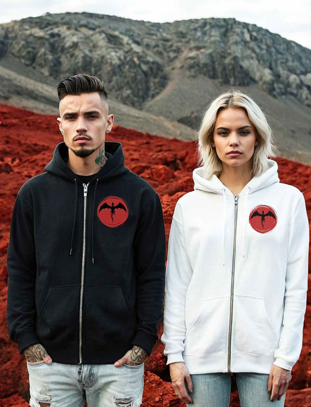 Ein männliches und ein weibliches Model posieren nebeneinander in 'Saga of the Dragon' Zip Hoodies, Schwarz und Weiß, vor einem Hintergrund roter Erde und ferner Berge, ein Bild kraftvoller Eleganz.