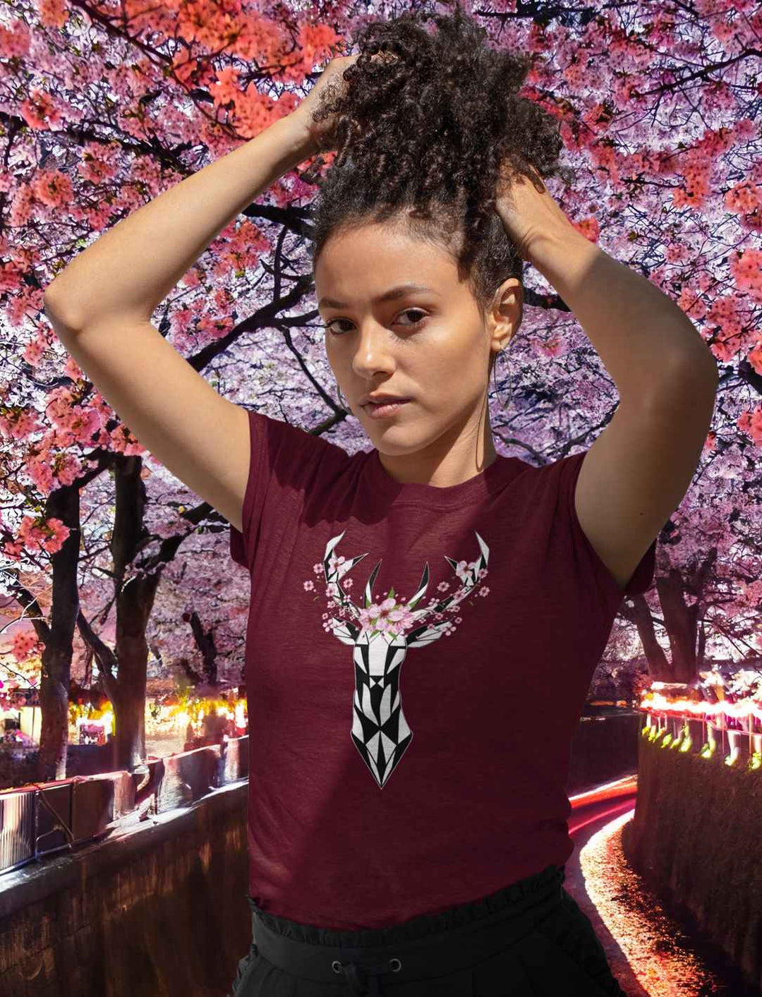 Eine junge Frau in einem burgunderfarbenen "Sakura Deer" T-Shirt, die das künstlerische Hirschmotiv umgeben von Kirschblüten zeigt, inspiriert von japanischen Frühlingslandschaften.