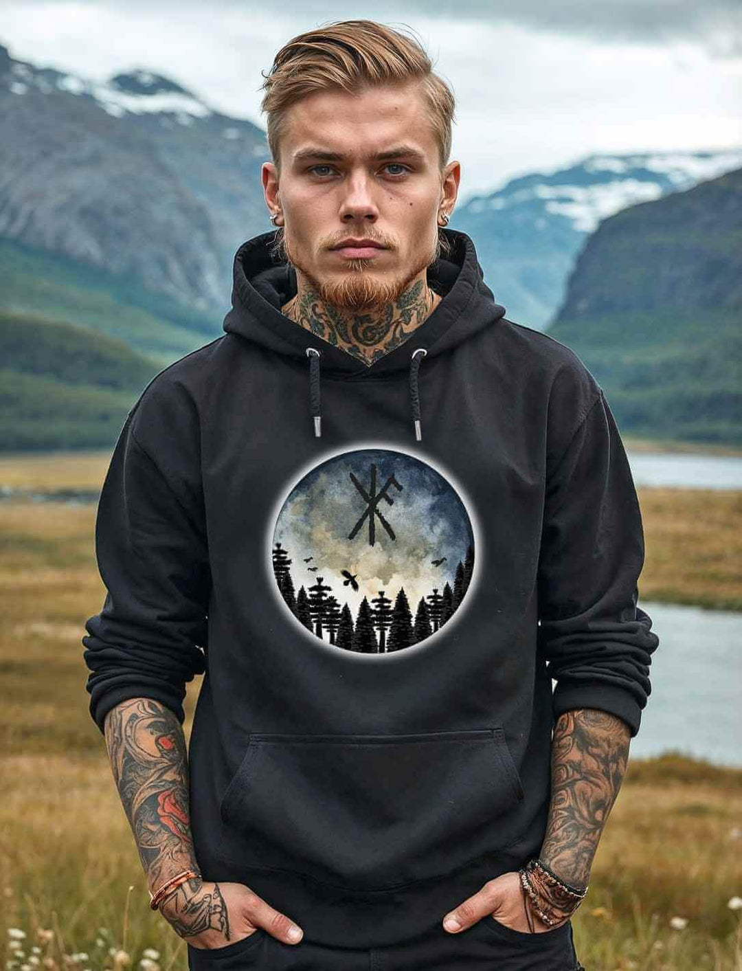 Ein Mann trägt den Unisex Organic Hoodie “Schutzrune der ewigen Stärke” mit einem mystischen Design, das eine Binderune, einen Wald, Vögel und einen stimmungsvollen Himmel zeigt. Fotografiert vor einer beeindruckenden Landschaft mit Bergen und Flüssen im Hintergrund.