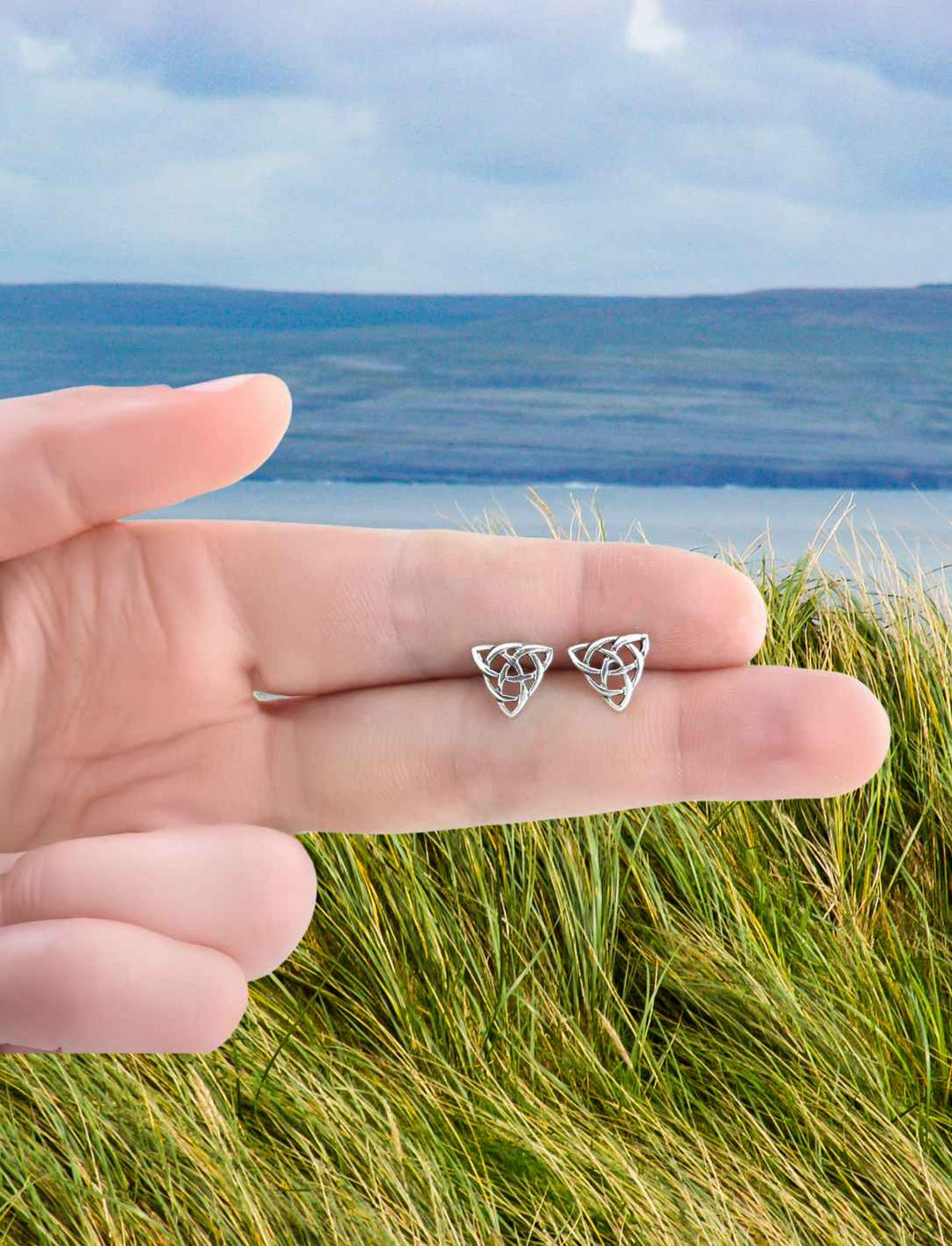 Silberne Ohrstecker der Triquetra aus 925er Sterling Silber, gehalten in einer Hand, im Hintergrund Grass und Meer.