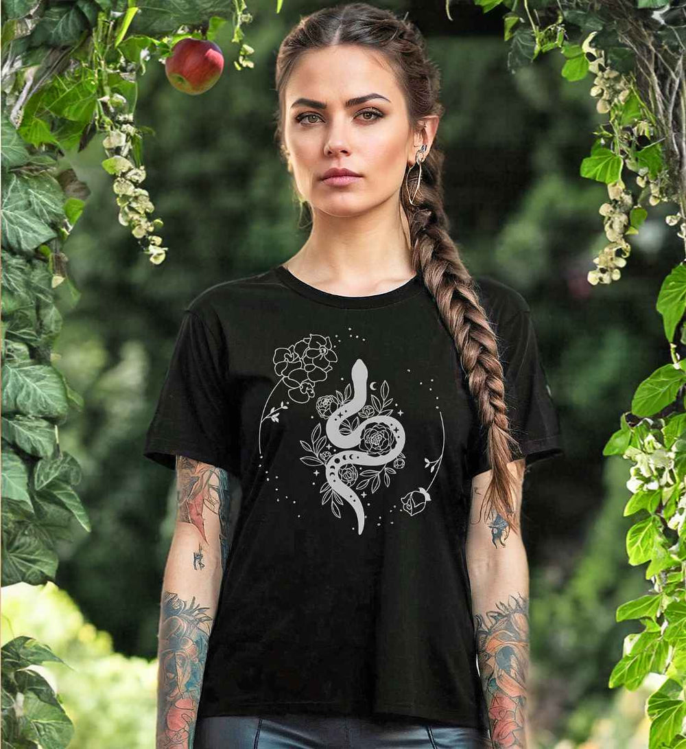 Junge Frau mit Zopffrisur trägt das 'Snake of Wisdom' T-Shirt in Schwarz, das mit einem weißen Schlangen- und Blumenmuster bedruckt ist, vor einem üppigen grünen Gartenhintergrund, der die Verbindung zur Natur unterstreicht