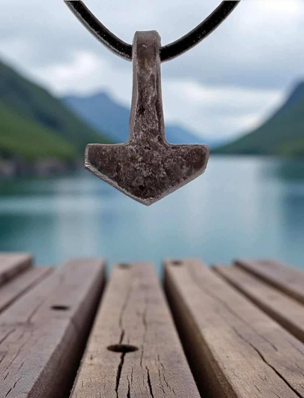 Thorshammer-Anhänger aus schwarzem Eisen hängt an einem Lederband frei über einem Holzsteg mit Blick auf einen See und umgebende Berge. Das Motiv vermittelt nordische Ruhe und Naturverbundenheit.