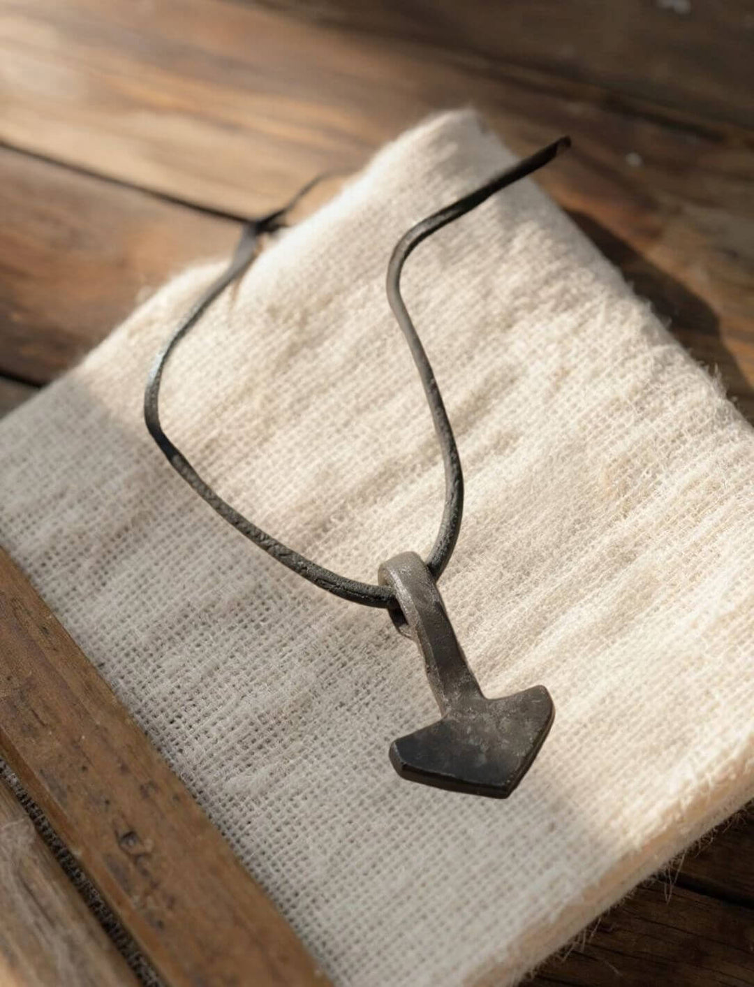 Thorshammer aus schwarzem Eisen mit Lederband liegt auf einem groben Stofftuch, das auf einer Holzfläche arrangiert ist. Das Licht fällt sanft auf das Amulett und hebt die Schmiedestruktur hervor.