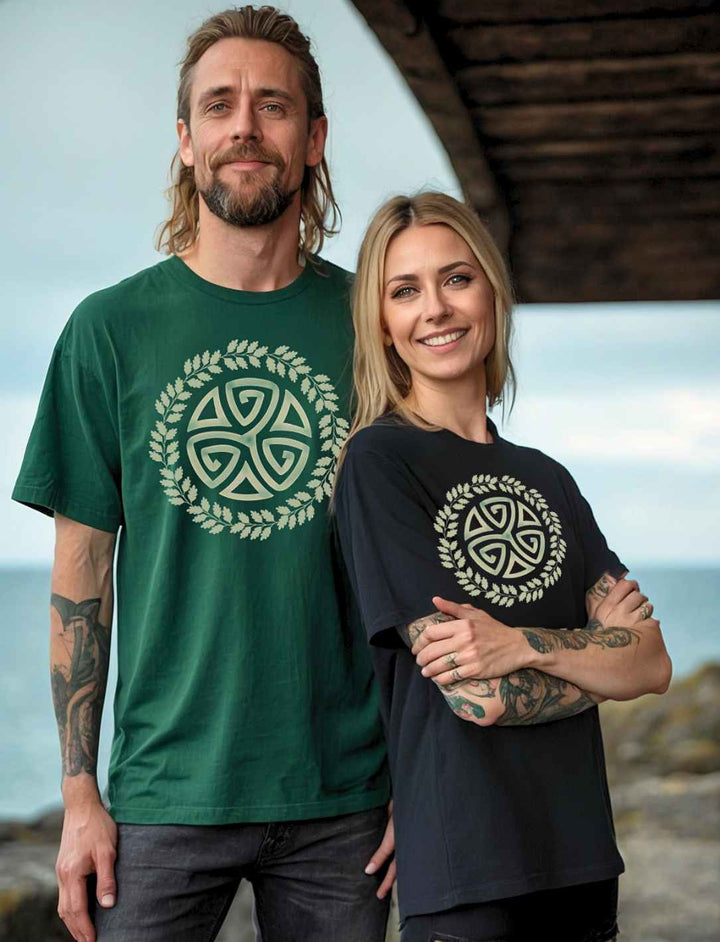 Ein Paar in "Triskele des Waldes" Heavy Oversized T-Shirts posiert vor einer Küste. Die Frau trägt das Design in Schwarz, der Mann in Glazed Green. Beide Shirts zeigen eine keltische Triskele, umgeben von einem Eichenblatt-Kranz, und verbinden Natur und Mythen mit modernem Stil.