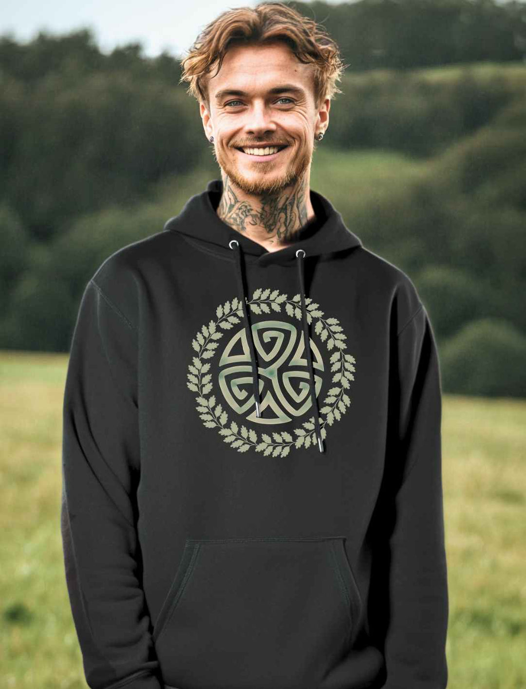 Junger Mann trägt das Triskele des Waldes Hoodie in schwarz. Im Hintergrund eine grüne Wiese und Wald.