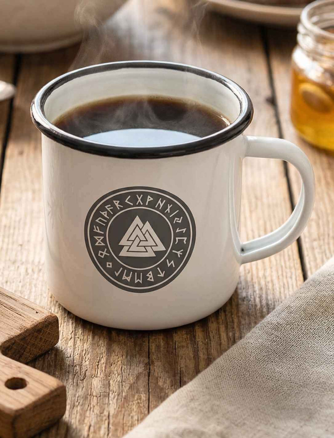 Valknut Emaille Tasse weiß mit schwarzem Rand und nordischem Odins Zeichen, gefüllt mit Kaffee auf rustikalem Holztisch
