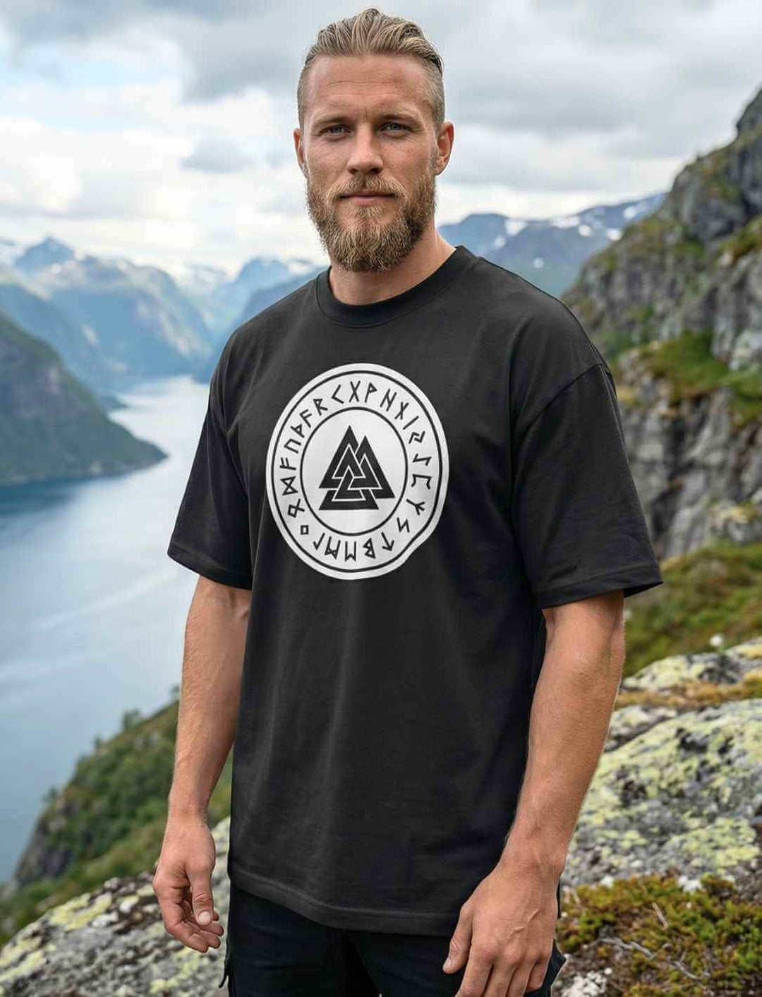  Mann trägt schwarzes Oversized Valknut T-Shirt mit nordischem Odin Symbol vor Fjordlandschaft