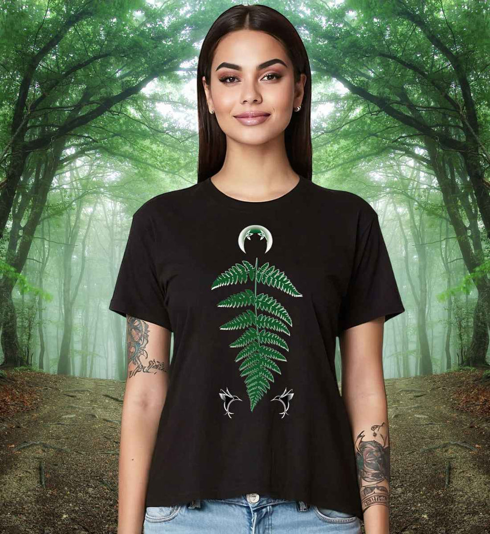 Frau mit langen dunklen Haaren trägt ein schwarzes Damen-T-Shirt 'Zauberhafte Naturwelt' mit grünem Farnblatt- und weißen Libellen-Motiv, vor einem Waldhintergrund posierend.
