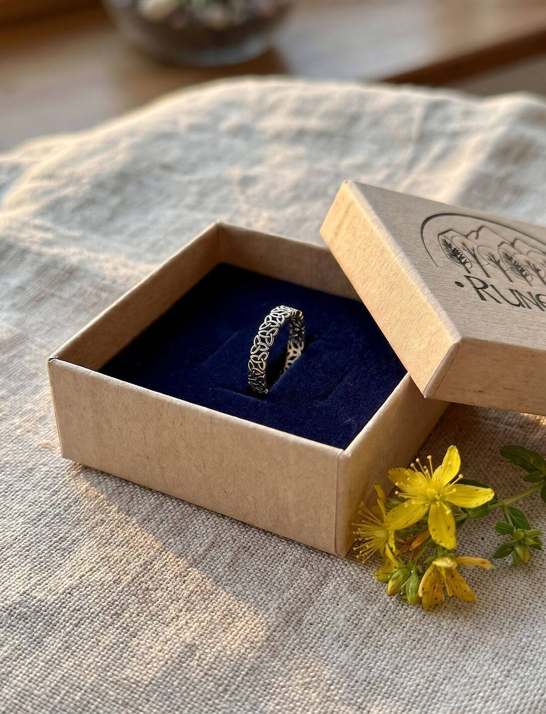 Triquetra Bandring in der Runental Geschenkbox aus Karton mit blauem Samt, daneben ein Zweig Johanniskraut