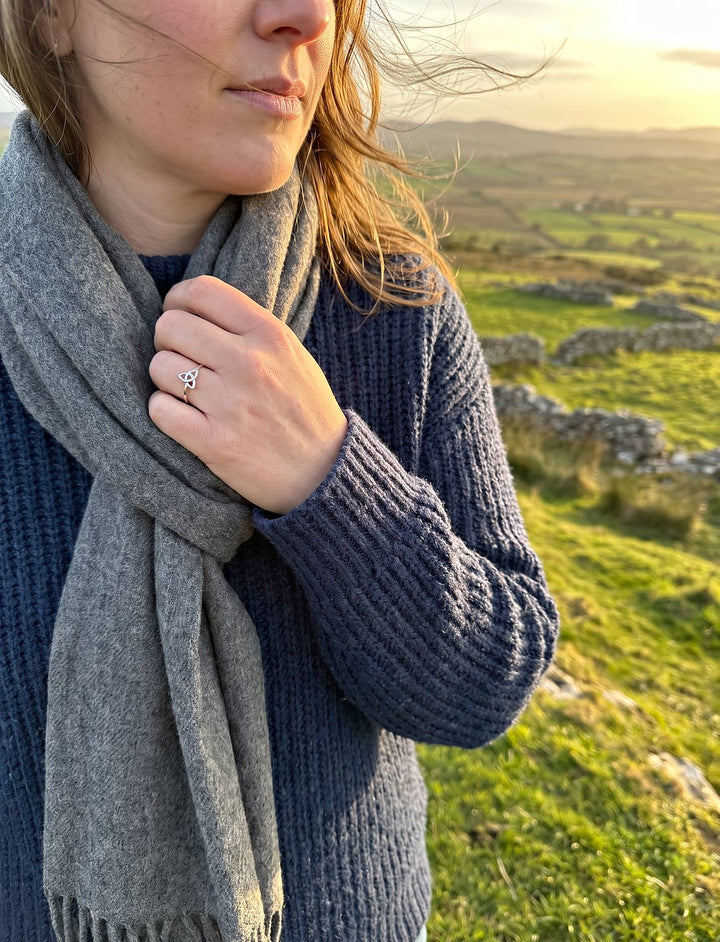 Frau im dunkelblauen Strickpullover trägt den Silberring mit Dreifaltigkeitsknoten und hält ihren grauen Schal im Wind