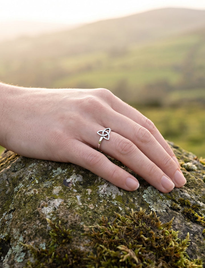 Schmaler Triquetra Silberring am Finger einer Frauenhand auf moosigem Fels, im Gegenlicht über einem weiten Tal