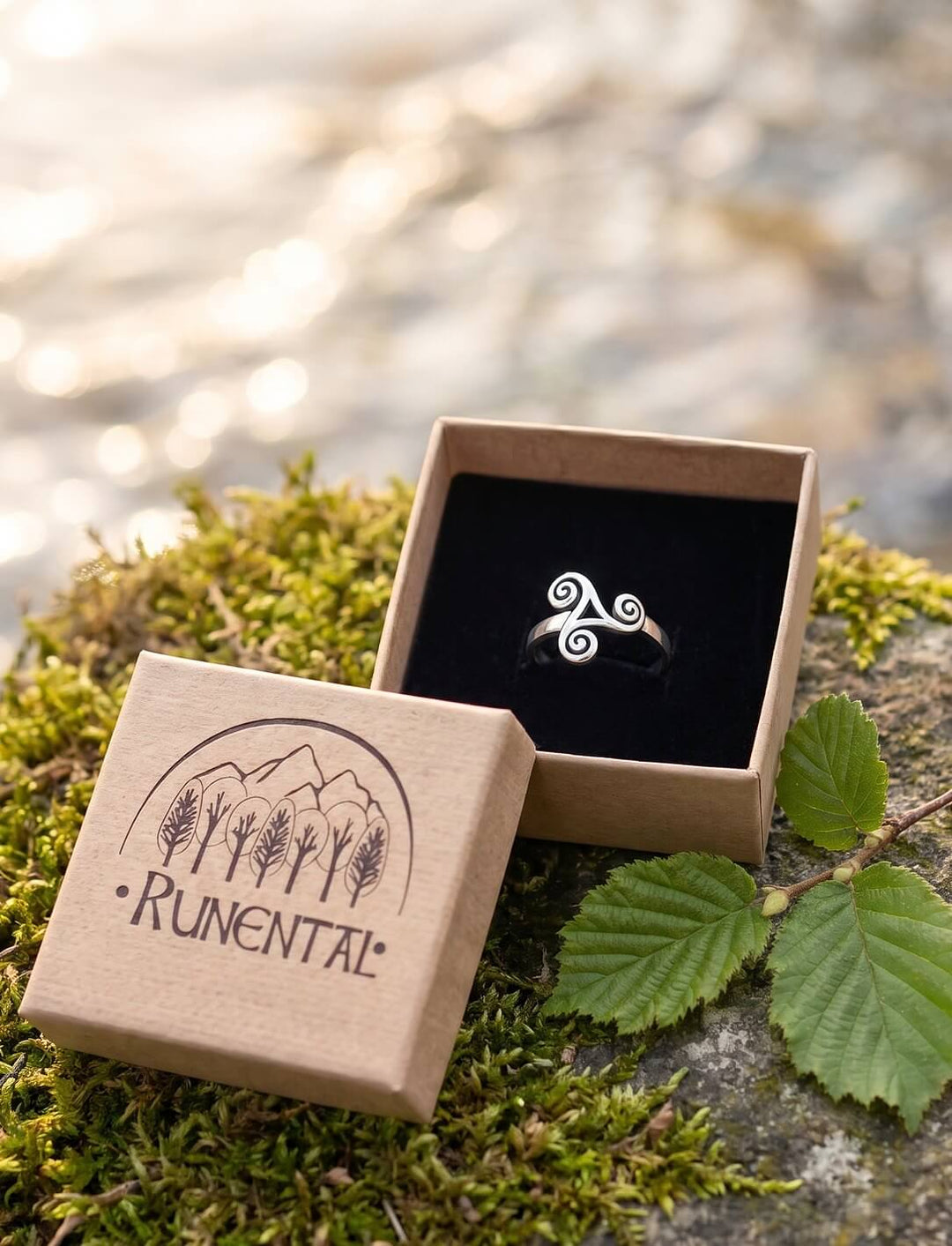 Triskele Ring in der Runental Geschenkbox aus Karton auf Moos, daneben ein Haselzweig vor glitzerndem Wasser
