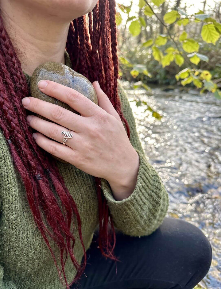 Frau mit roten Zöpfen und grünem Strickpullover hält einen Flusskiesel, am Finger ein Silberring mit dreifacher Spirale