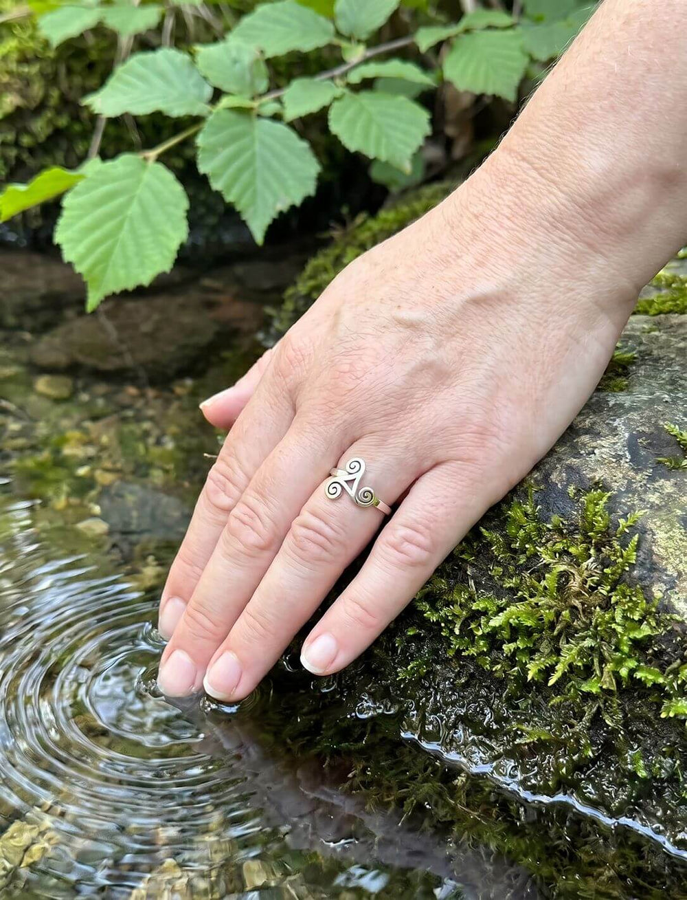Frauenhand mit keltischem Spiralring aus Silber am Ringfinger berührt das klare Wasser einer Quelle neben Moos