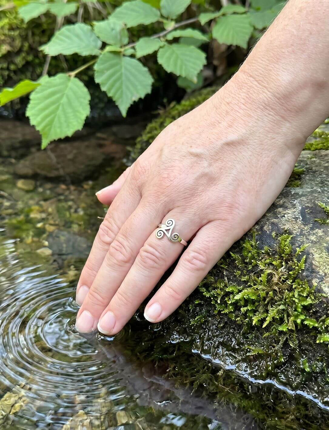 Frauenhand mit keltischem Spiralring aus Silber am Ringfinger berührt das klare Wasser einer Quelle neben Moos