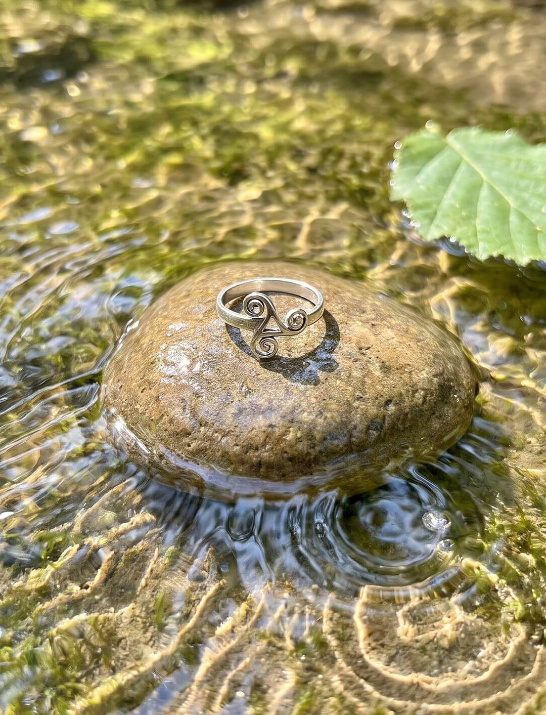 Silberring mit drei keltischen Spiralen liegt auf einem nassen Kieselstein im flachen Quellwasser mit Haselblatt