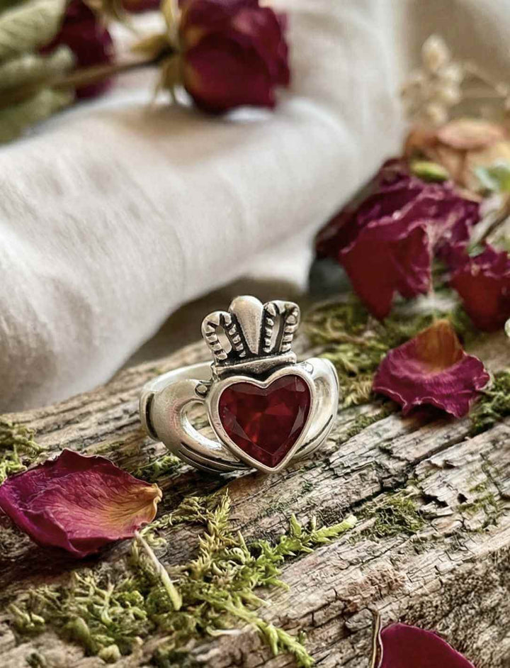 Claddagh Ring mit rotem Herzstein auf Holz mit Rosenblättern und Kerzenlicht in keltischer Umgebung