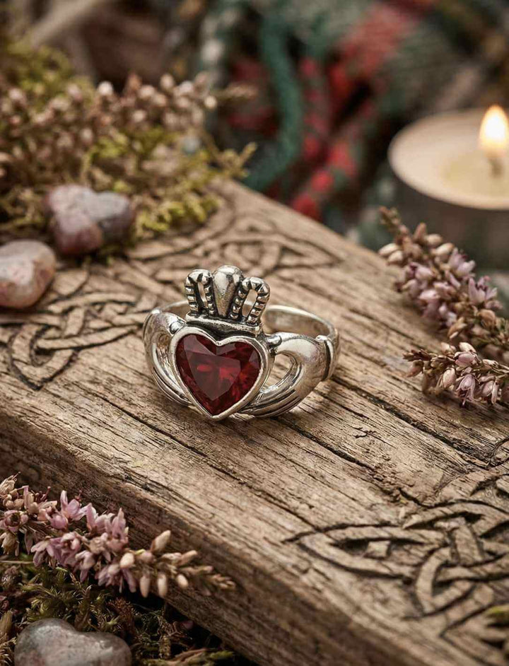 Claddagh Ring aus Silber mit rotem Zirkonia auf keltisch verzierter Holztafel im moosigen Wald