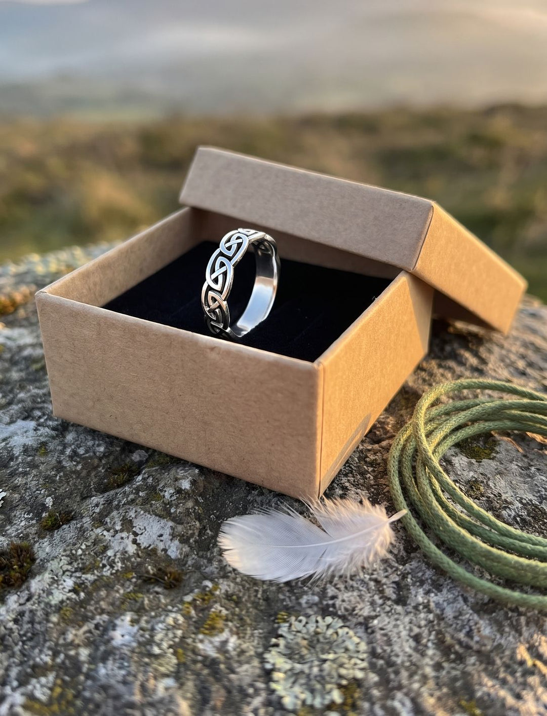 Keltischer Ehering aus Silber in der Runental Geschenkbox aus Pappe, daneben grüne Kordel und weiße Feder auf Moos