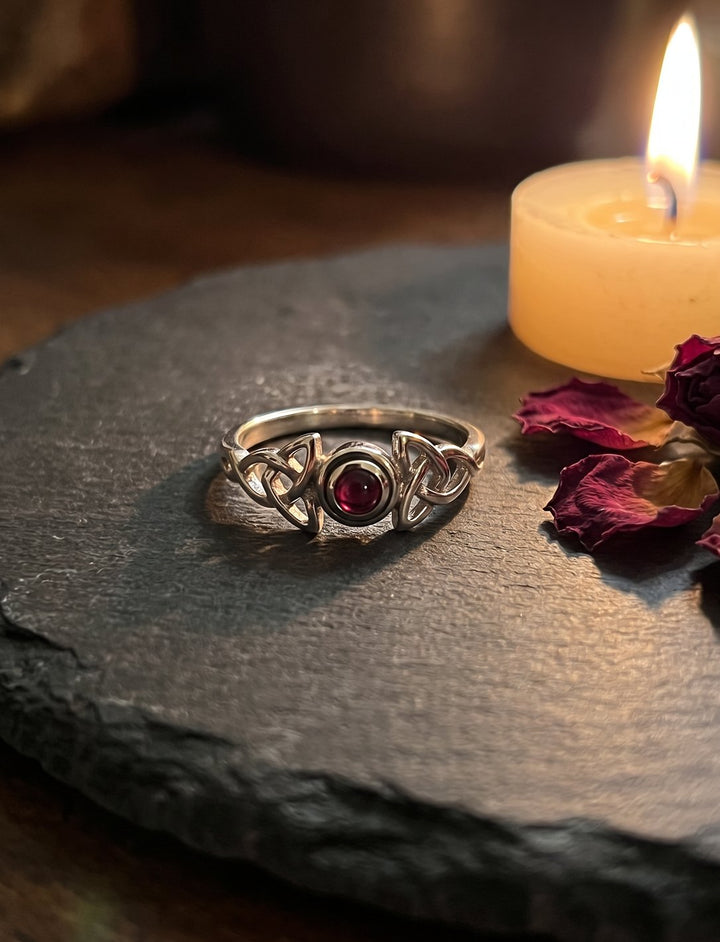 Silberring mit Dreiecksknoten und rundem Granat auf Schiefer neben Kerze und getrockneten Rosenblättern