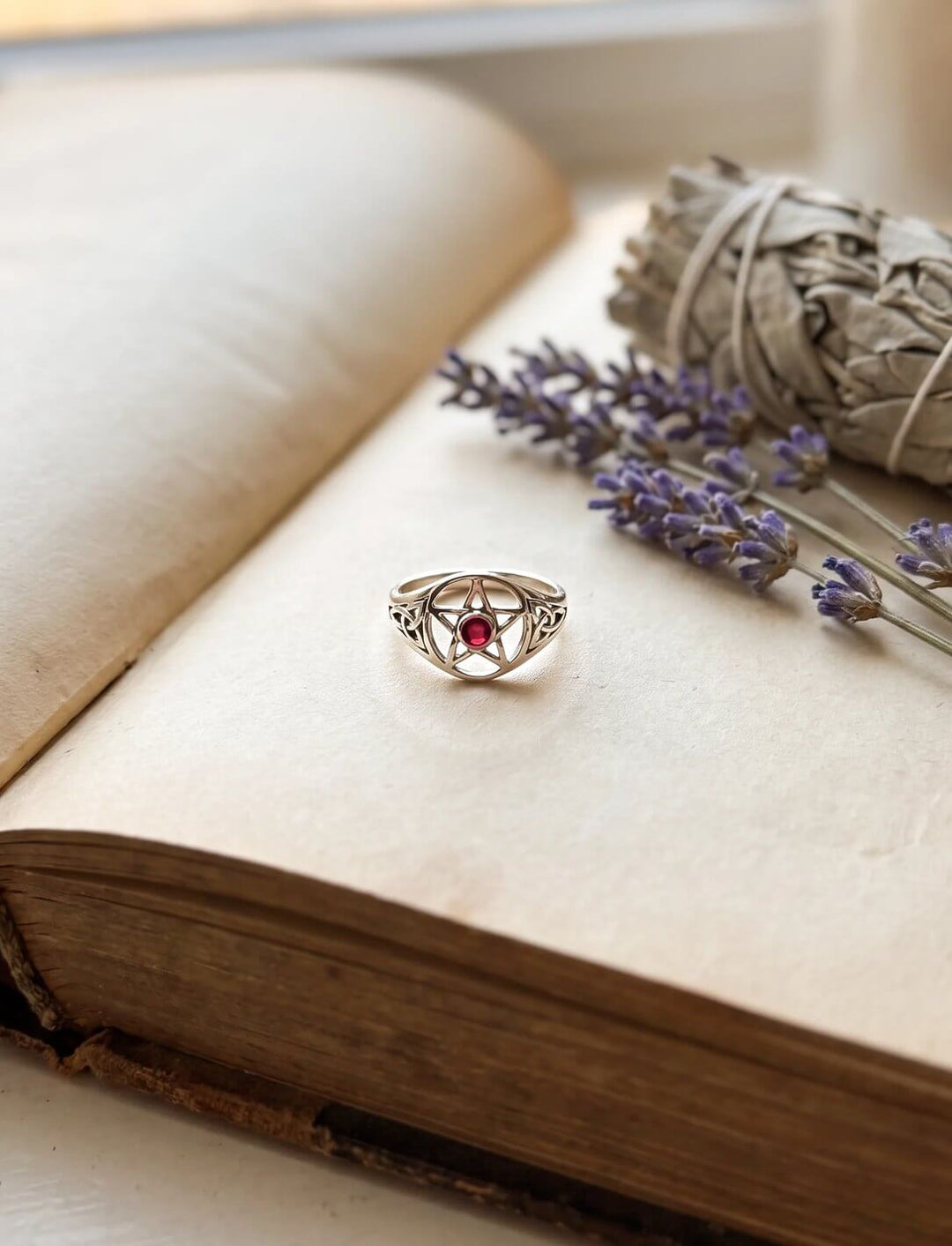 Keltischer Pentagramm Ring mit Triquetras und Granat liegt auf einem alten Buch neben Lavendel und Salbei