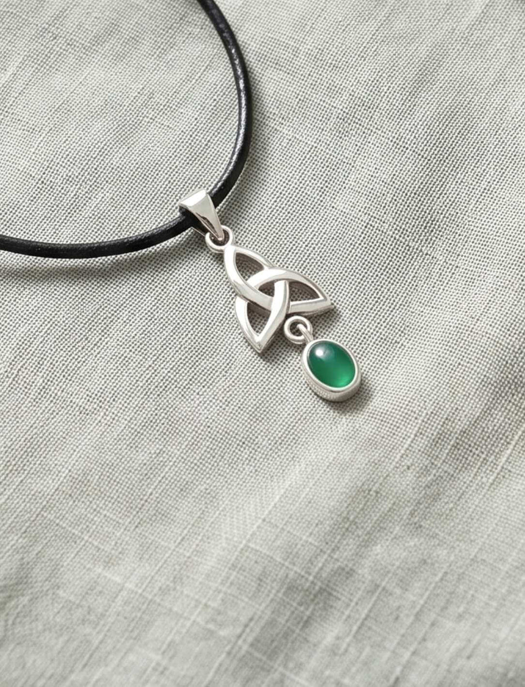 Keltischer Triquetra Anhänger mit grünem Achat am Lederband neben hellen Kieseln und Farnen auf Leinen