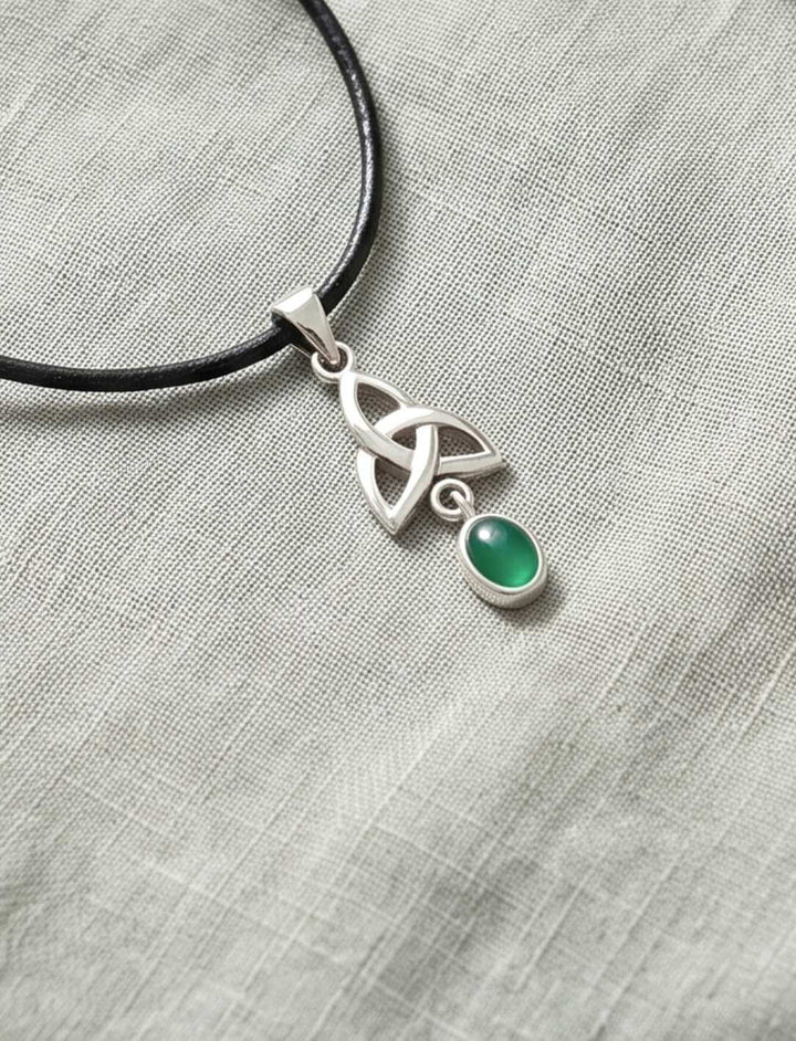 Keltischer Triquetra Anhänger mit grünem Achat am Lederband neben hellen Kieseln und Farnen auf Leinen