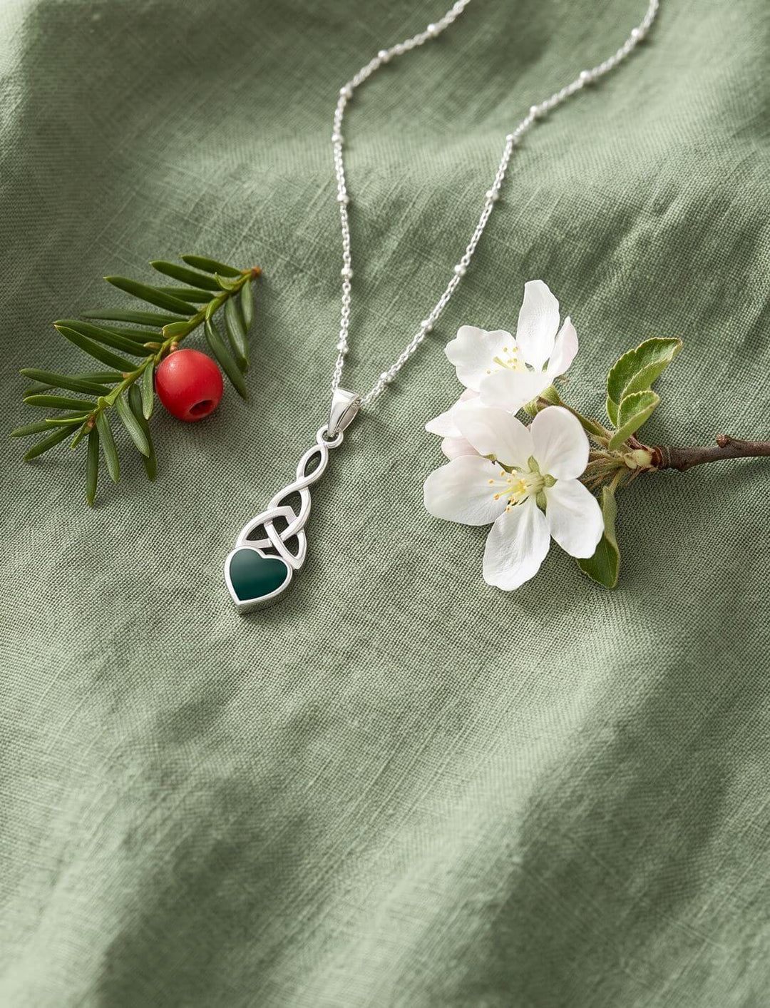 Triquetra Anhänger mit grünem Achat-Herz an Himmelsperlen-Kette auf grünem Leinen neben Eibe und Apfelblüte