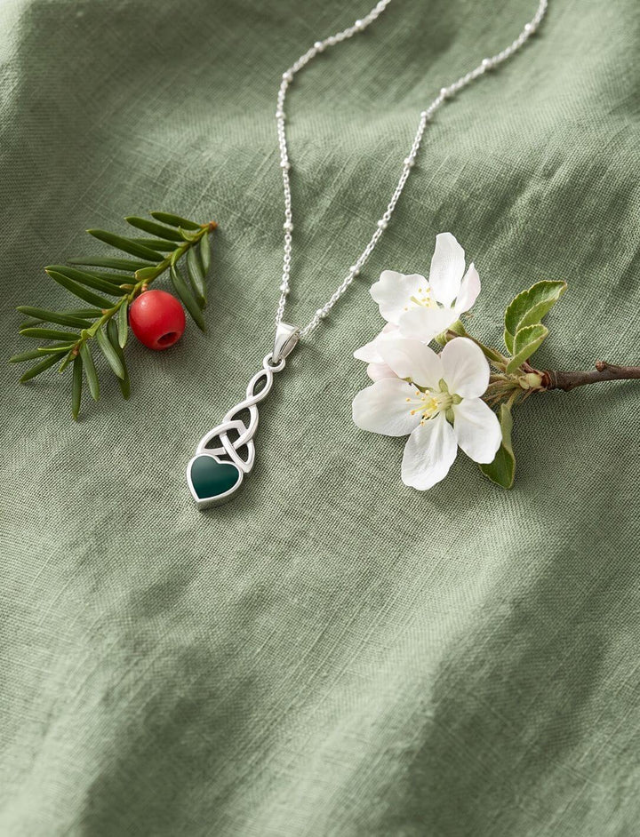 Triquetra Anhänger mit grünem Achat-Herz an Himmelsperlen-Kette auf grünem Leinen neben Eibe und Apfelblüte