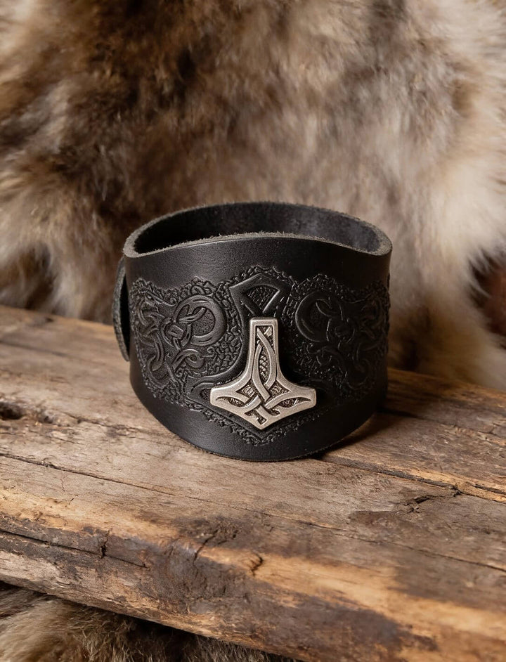 Schwarzes Lederarmband mit silberfarbenem Mjölnir-Beschlag und geprägten Tierornamenten auf einem Holzbrett vor Fell