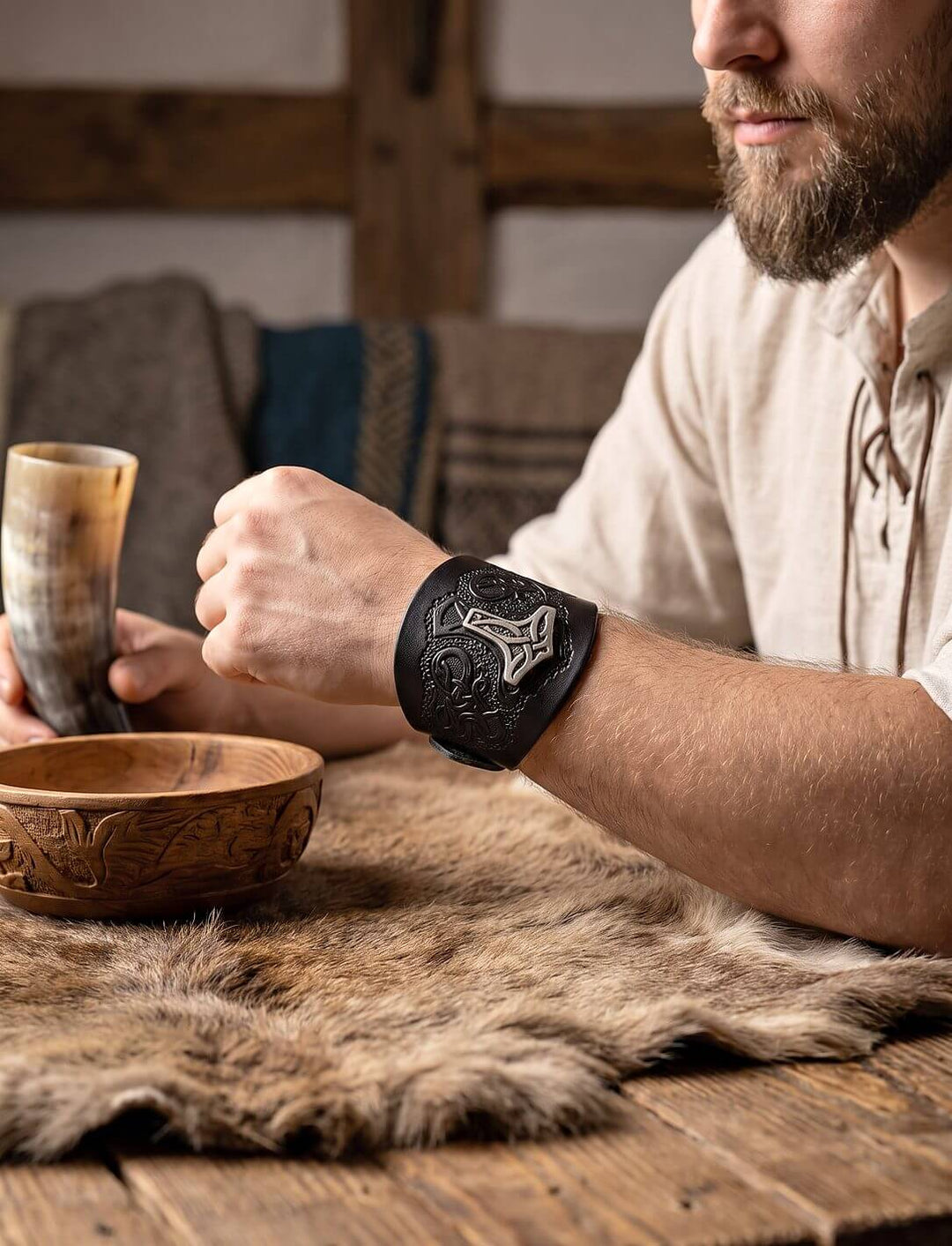 Mann im Leinenhemd trägt das schwarze Wikinger Armband mit Thorshammer am Handgelenk, daneben Trinkhorn und Holzschale