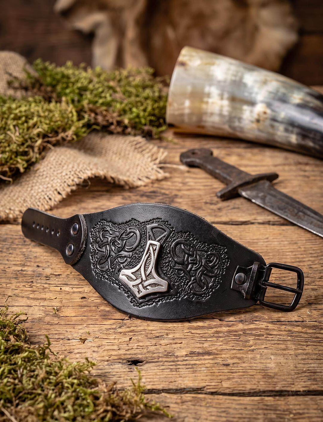 Járngreipr: schwarzes Thors Hammer Armband aus geprägtem Leder mit Dornschnalle, liegend neben Trinkhorn und Moos