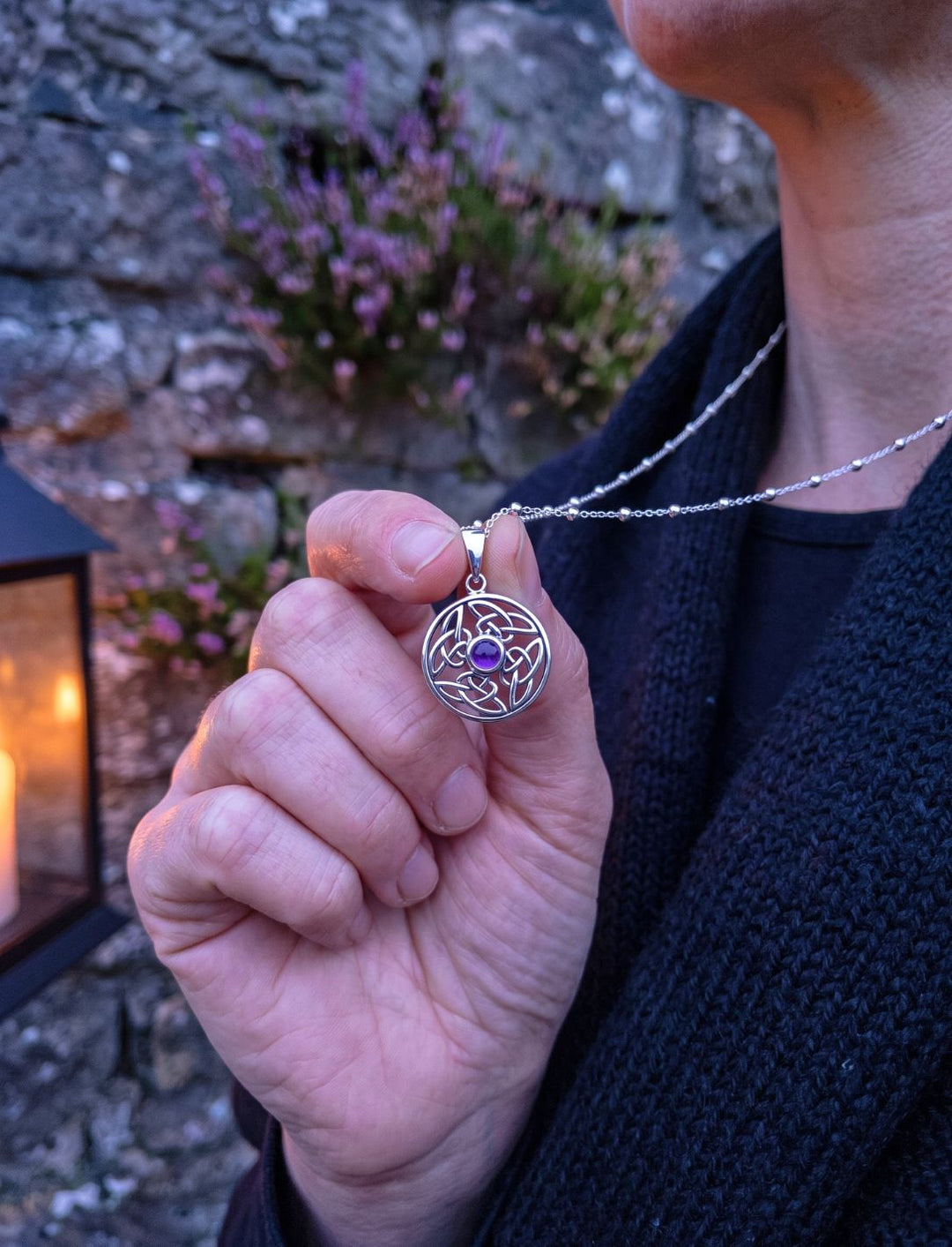 Hand hält Amethyst Anhänger an Silberperlenkette vor Steinmauer mit Heide und Laterne in der Dämmerung