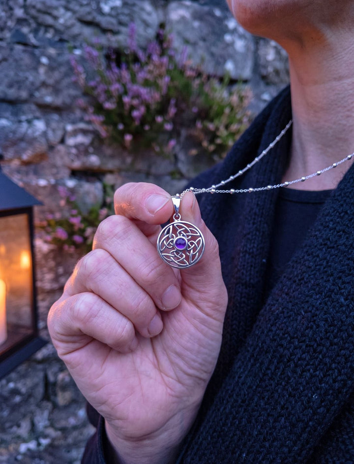 Hand hält Amethyst Anhänger an Silberperlenkette vor Steinmauer mit Heide und Laterne in der Dämmerung