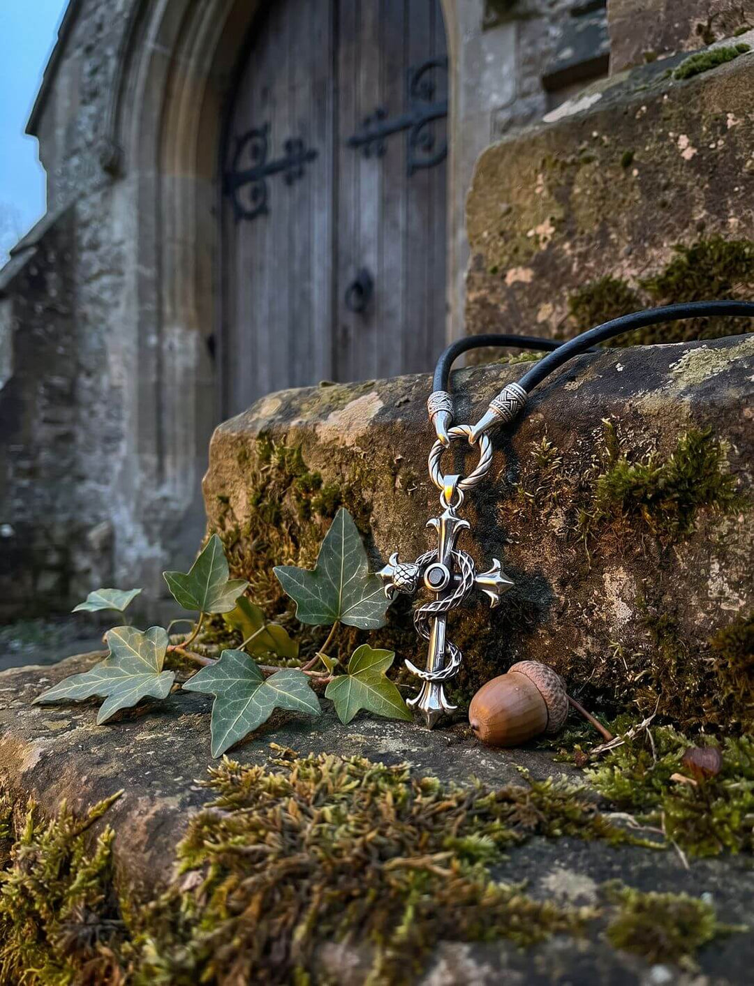 Kreuzanhänger mit Lilienenden am Band des Hüters auf moosiger Steinmauer vor alter Kirchentür, mit Efeu und Eichel