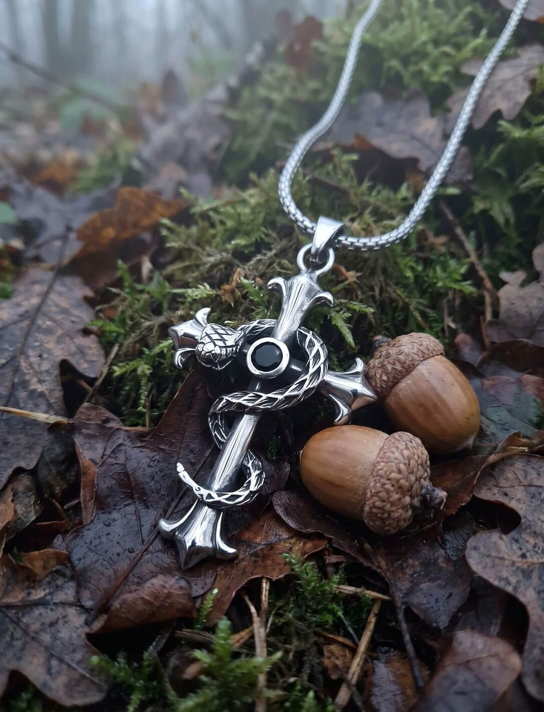 Lilienkreuz mit gewundener Schlange an Edelstahl-Schlangenkette auf Moos und Herbstlaub zwischen zwei Eicheln