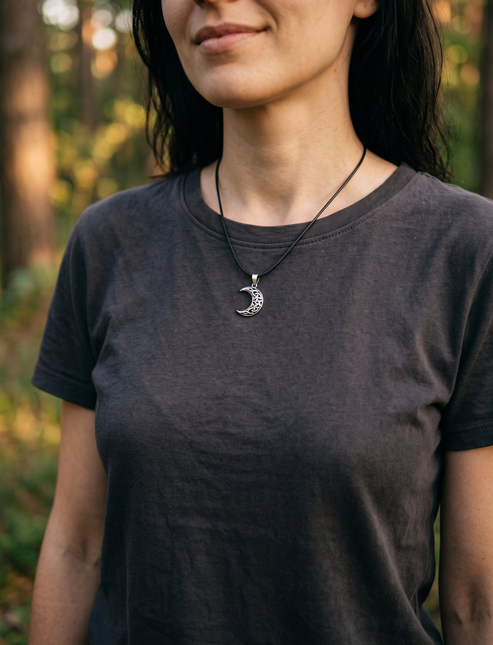 Frau im dunklen Shirt trägt die schmale Mondsichel mit keltischem Knoten an Lederkordel, warmes Waldlicht dahinter