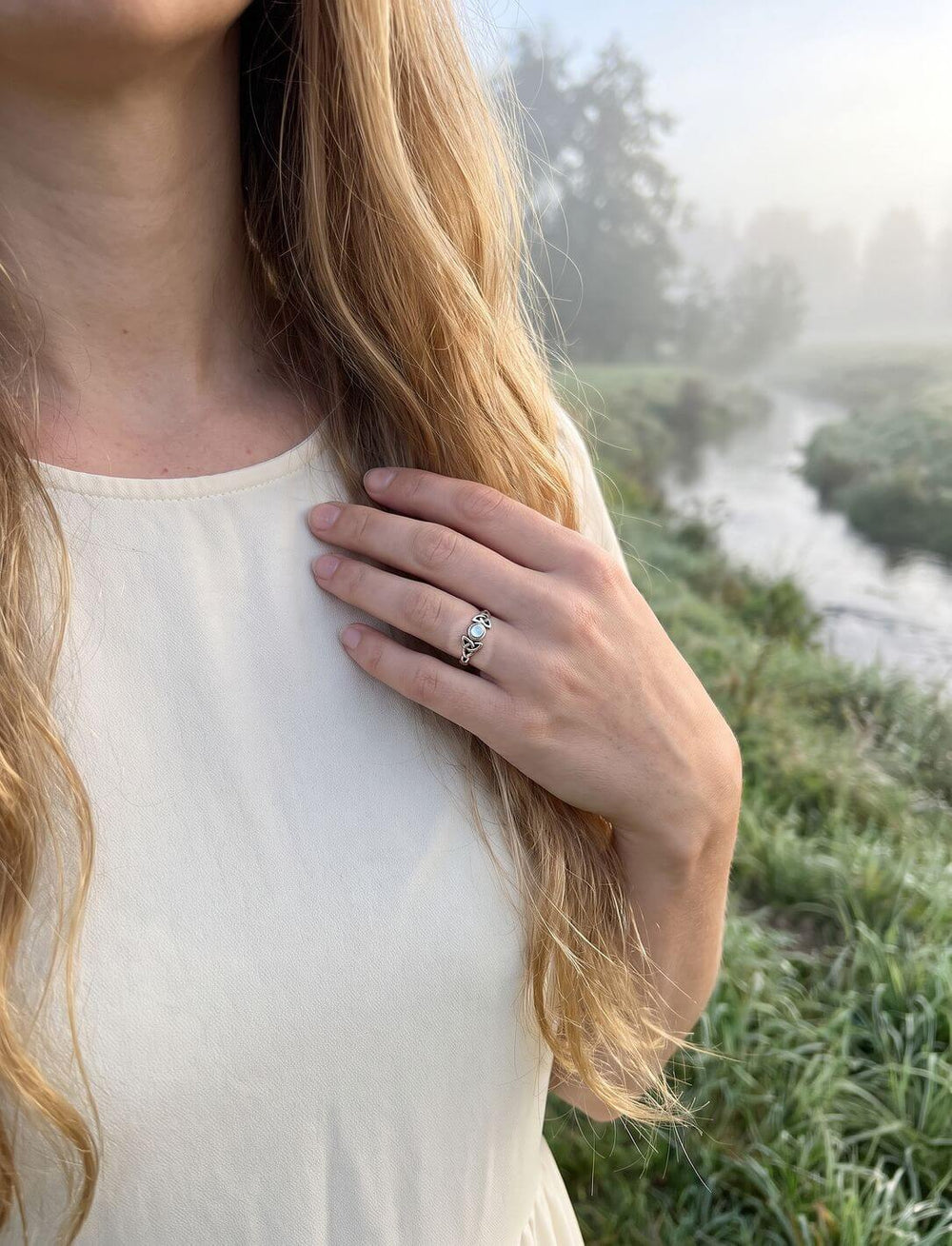 Frau im cremeweißen Kleid trägt den keltischen Knotenring mit Regenbogenmondstein an einem Bach im Morgennebel